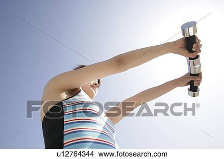 Picture - Low angle view of a young woman holding two dumbbells. Fotosearch
