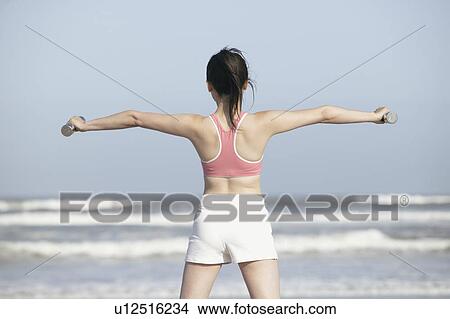 Rear view of a woman stretching her arms at the seashore with dumbbells in her hands View Large Photo Image Picture - Rear view of a woman stretching her arms at the seashore with dumbbells in her hands. Fotosearch