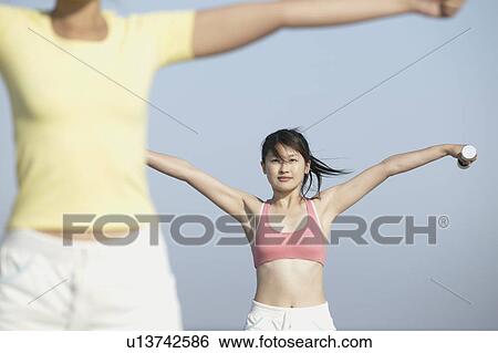 Stock Photograph - Two women stretch their arms as they hold dumbbells during exercise. Fotosearch