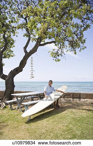 Picture - Mature man sitting on bench with surfboard. Fotosearch