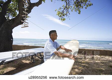 Stock Photograph - Mature man sitting on bench with surfboard. Fotosearch
