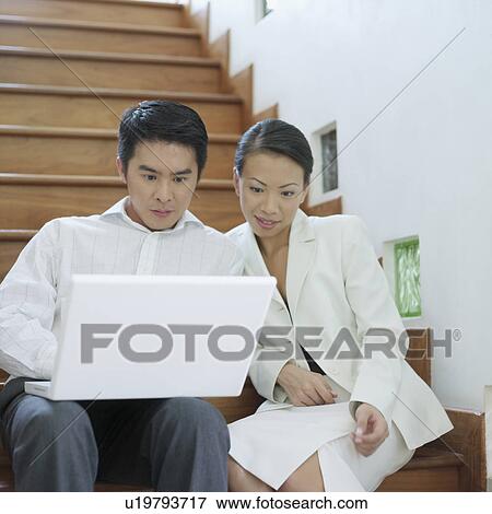 Stock Photo - Businessman and businesswoman sitting on a staircase and looking at a laptop. Fotosearch