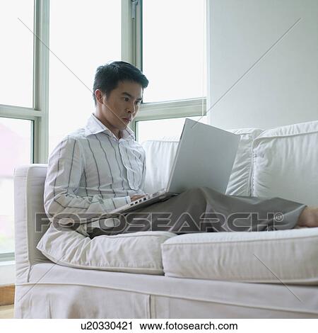 Young man sitting on a couch using a laptop View Large Photo Image Stock Image - Young man sitting on a couch using a laptop. Fotosearch