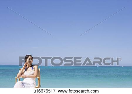 Stock Photo - A smiling young woman sitting on chair at beach. Fotosearch