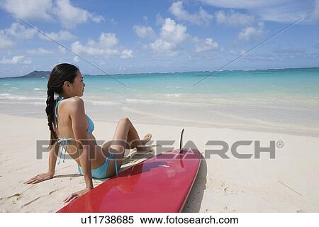 Stock Photography - A woman sitting on beach with surfboard. Fotosearch