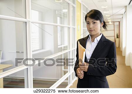 A businesswoman holding a file stands in the passage of the office View Large Photo Image Stock Photography - A businesswoman holding a file stands in the passage of the office. Fotosearch