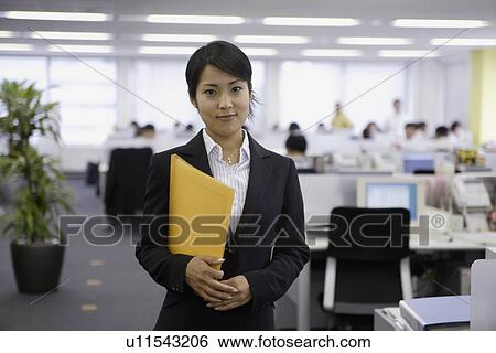 Stock Photograph - A professional woman holding a file smiles at the camera in the office. Fotosearch