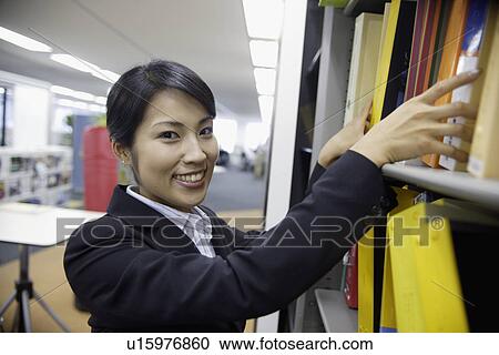 Stock Image - A woman smiles at the camera while finding a file from the shelf. Fotosearch