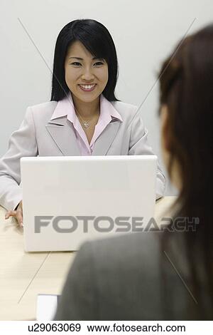 Stock Photo - A woman working on her laptop smiles at her colleague. Fotosearch