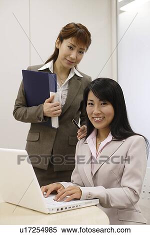 Stock Photography - Two women faces the camera as one of them works on the laptop. Fotosearch