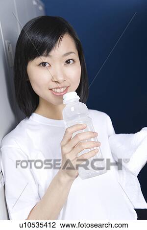 Stock Image - Portrait of a teenage girl in gym cloth smiling and holding a bottle of water in locker room. Fotosearch