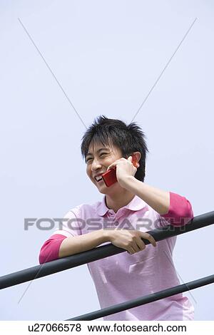 Stock Photo - Man leaning on the railing and using mobile phone under the blue sky, low angle view, blue background, Japan. Fotosearch