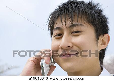 Stock Photograph - Portrait of a man using mobile phone under the blue sky, smiling and looking at camera, front view, close up, Japan. Fotosearch