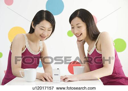 Stock Photo - Two Young Twin Sisters Sitting at Table, Using a Mobile Phone, Front View. Fotosearch