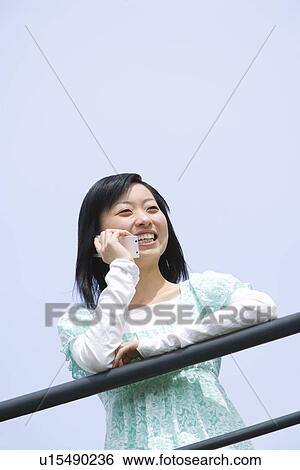 Stock Photograph - Woman leaning on the railing and using mobile phone under the blue sky, low angle view, blue background, Japan. Fotosearch