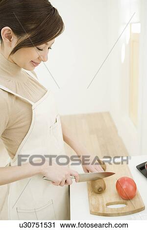 Stock Image - A woman cutting a kiwi fruit in the kitchen, side view, high angle view. Fotosearch