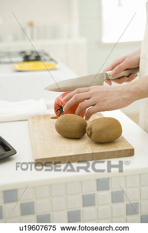 Stock Photography - A woman cutting a tomato in the kitchen, side view, differential focus. Fotosearch