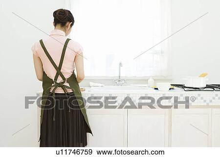 Stock Photo - A woman standing in the kitchen, rear view. Fotosearch