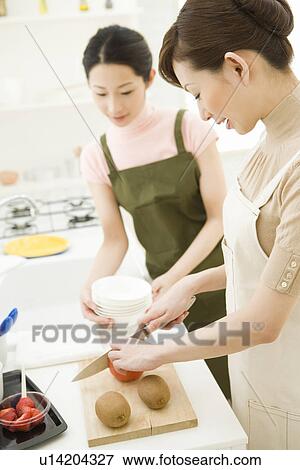 Stock Photo - Two women in the kitchen, one cutting fruit, front view, side view, differential focus. Fotosearch