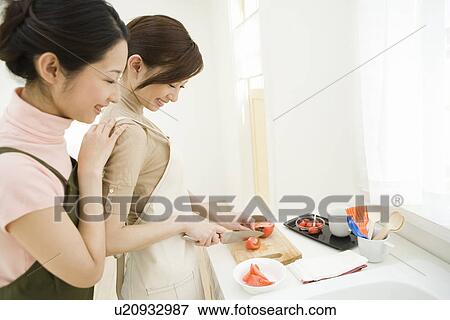 Stock Photo - Two women in the kitchen, one cutting tomatoes, side view, high angle view. Fotosearch