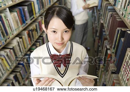 Stock Photo - Portrait of a teenage girl holding a book in library, smiling and looking at camera, boy in background. Fotosearch