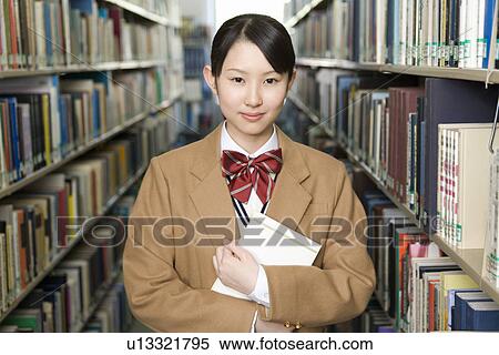 Stock Photography - Portrait of a teenage girl holding a book in library, smiling and looking at camera. Fotosearch