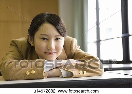 Stock Image - Portrait of teenage girl sitting and leaning on desk in classroom, smiling and looking at camera. Fotosearch
