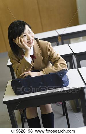Stock Photo - Teenage girl smiling and sitting at desk in classroom, resting chin in hand. Fotosearch