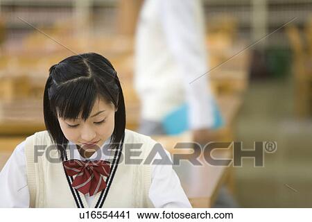 Stock Image - Teenage girl studying in library, boy walking in background, differential focus. Fotosearch