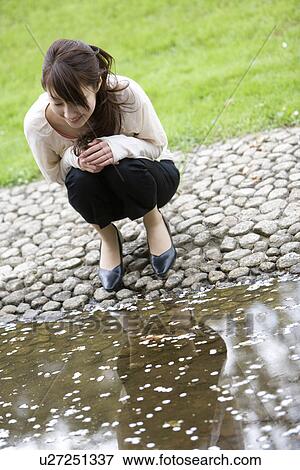 Stock Photo - A Young Woman Looking Down Petals of Cherry blossom Floating on Water, Front View. Fotosearch