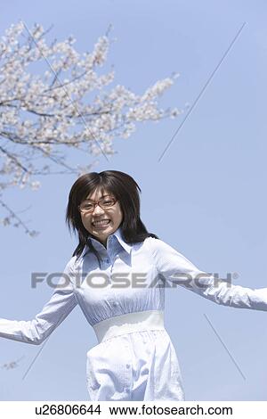 Picture - Portrait of a woman jumping under the cherry tree, smiling and looking at camera, front view, blue background, Japan. Fotosearch