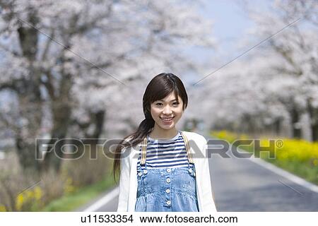 Picture - Portrait of a woman on the road lined with cherry trees, smiling and looking at camera, front view, Japan. Fotosearch