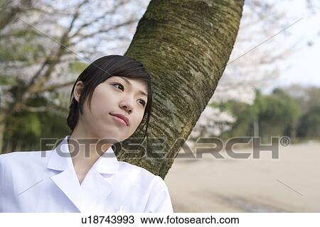 Stock Image - Woman in lab coat leaning on a tree trunk, front view, Japan. Fotosearch