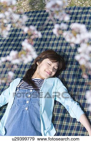 Stock Photo - Woman lying on a leisure sheet under the cherry tree, closing eyes, high angle view, differential focus, Japan. Fotosearch