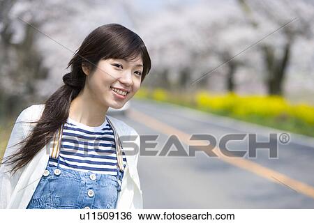 Stock Photograph - Woman standing on the side of a road lined with cherry trees, front view, Japan. Fotosearch