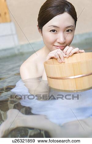 Stock Photo - Portrait of a woman in hot tub holding a wooden tub, looking at camera, front view, Japan. Fotosearch