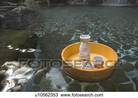 Stock Image - Sake bottle and cups in a wooden tub floating on a Japanese public bath, hot spring, high angle view, Japan. Fotosearch