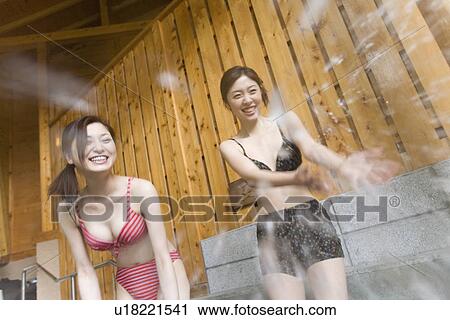 Stock Image - Two women splashing water in hot tub, in bathing suit, smiling, front view, Japan. Fotosearch