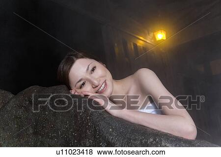 Stock Photo - Woman in hot tub leaning on the rock, smiling, front view, Japan. Fotosearch