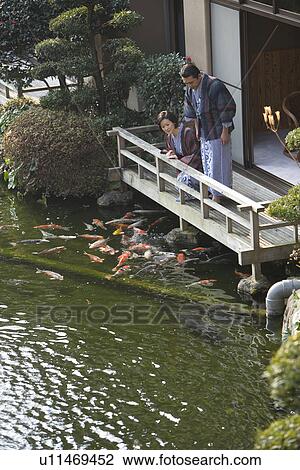 Stock Image - Mature Adult Couple Looking at Carp from the Wooden Balcony, Side View, High Angle View, Full Length, Three Quarter Length. Fotosearch