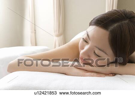 Stock Image - Woman lying on bed in beauty salon, closing eyes, front view, Japan. Fotosearch