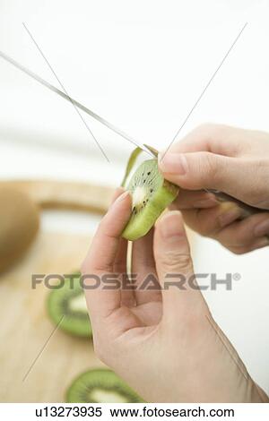 Stock Photography - A woman peeling a slice of kiwi fruit in the kitchen, high angle view, close up, differential focus. Fotosearch