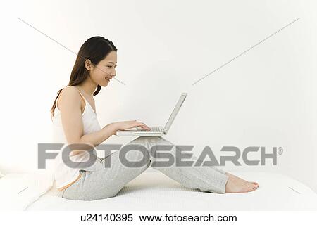 A woman sitting on a bed and using a laptop, side view, white background View Large Photo Image Stock Photography - A woman sitting on a bed and using a laptop, side view, white background. Fotosearch