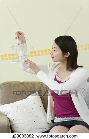 Stock Image - Woman sitting on couch and holding a birdcage, front view. Fotosearch