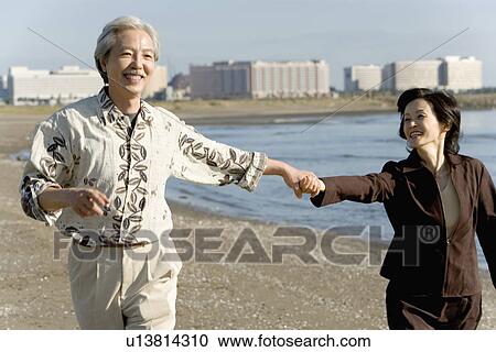 Stock Image - A Mature Couple Walking Hand in Hand on a Beach, Front View, Three Quarter Length. Fotosearch