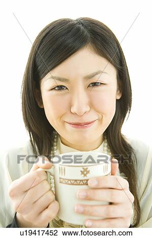 Stock Image - Portrait of a woman holding a mug, smiling and looking at camera, front view, white background. Fotosearch
