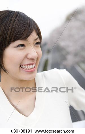 Stock Image - Portrait of a woman smiling, road lined with cherry trees in the background, front view, Japan. Fotosearch