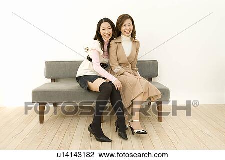 Stock Image - Portrait of two women smiling and sitting on the couch, looking at camera, front view. Fotosearch
