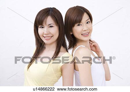 Stock Image - Two young women, back to back, smiling, front view. Fotosearch