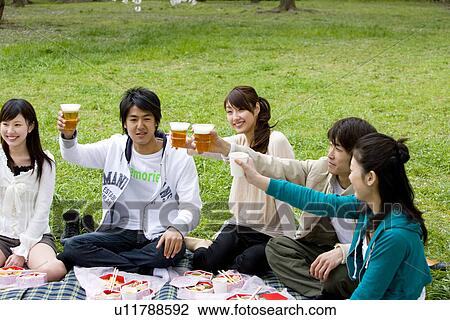 Stock Image - Five young people toasting with glasses, enjoying lunch on lawn, front view, side view, Japan. Fotosearch